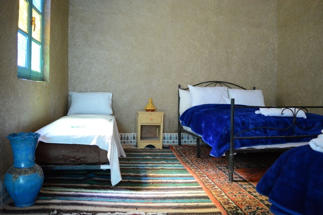 A room with two beds, a small table with a lamp, a blue vase, and a patterned rug.