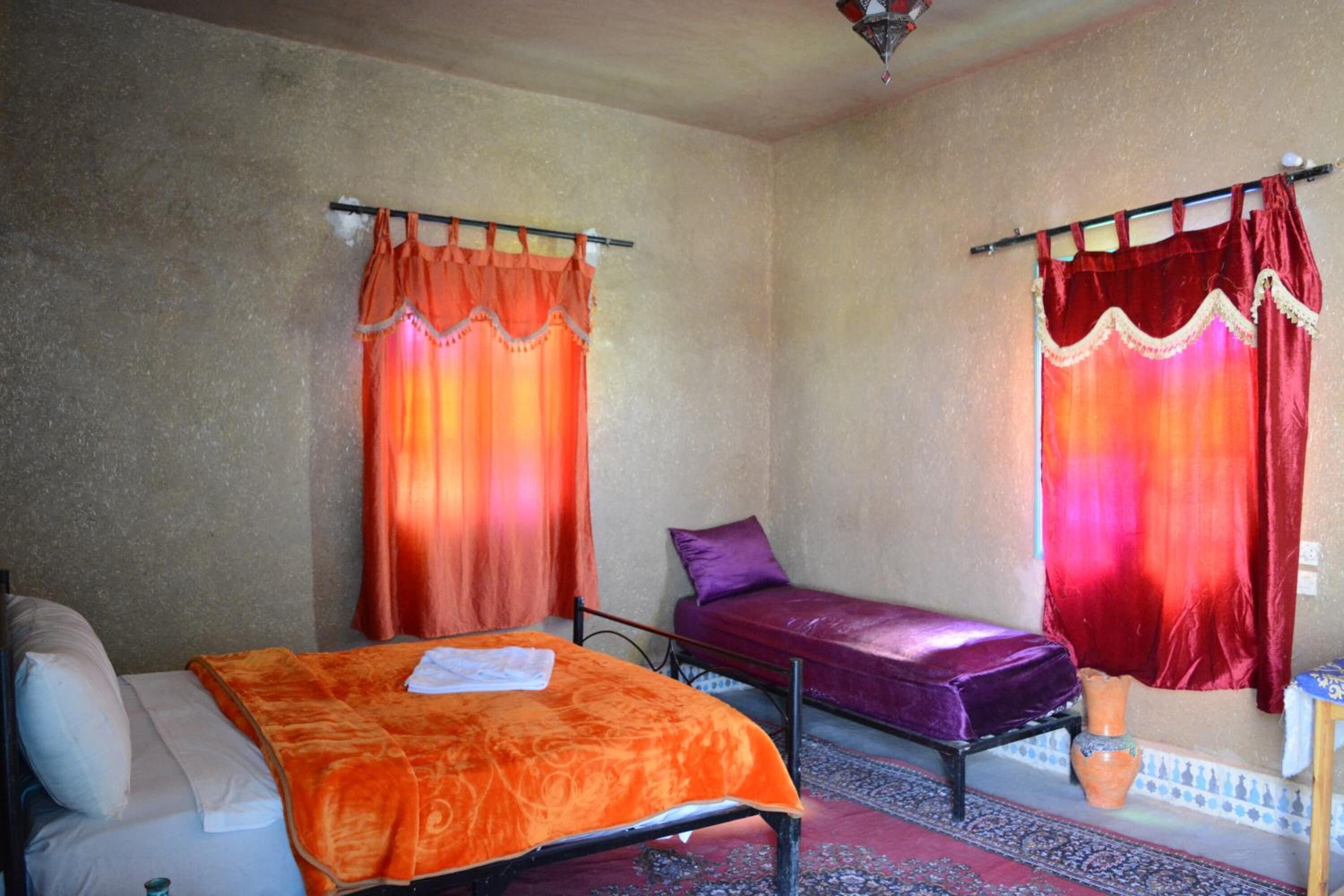 A room with two beds, one with an orange bedspread and the other with a purple bedspread. There is a small table with a vase on it.