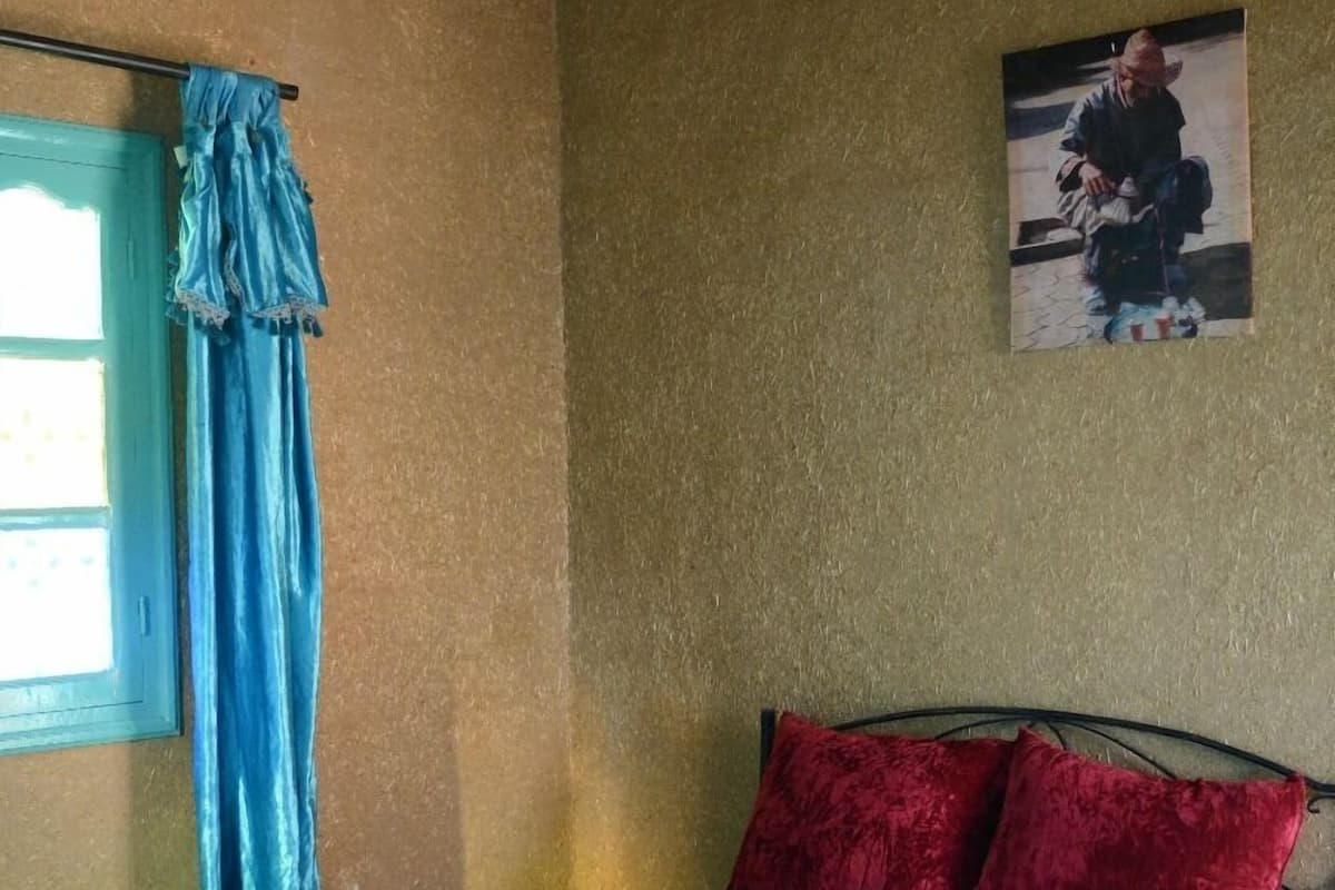 Un lit simple avec un cadre en métal, un matelas blanc et des oreillers rouges. Il y a un rideau bleu à la fenêtre et une ampoule qui pend au plafond.
