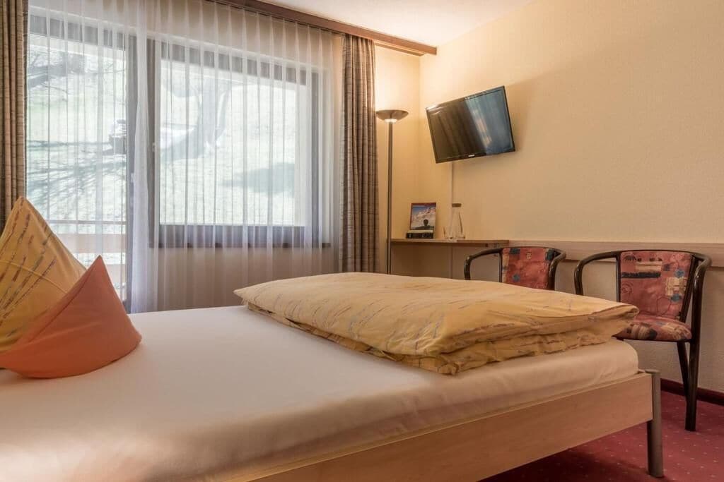 A hotel room with a bed, a TV mounted on the wall, a desk with a chair, and a window with curtains.