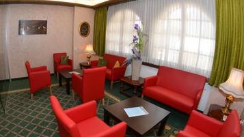 Habitación de hotel con un sofá rojo, dos sillones rojos, una mesa de centro y una mesita auxiliar. Se ve una ventana con cortinas verdes y un cuadro en la pared.