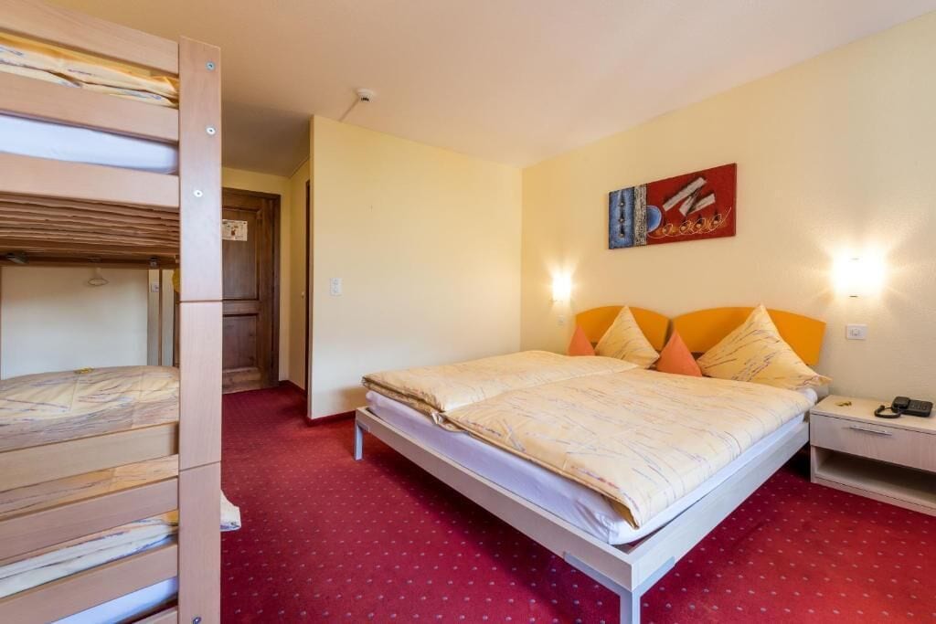 Una habitación de hotel con literas, una cama individual con almohadas naranjas, una mesita de noche con un teléfono y un cuadro en la pared.