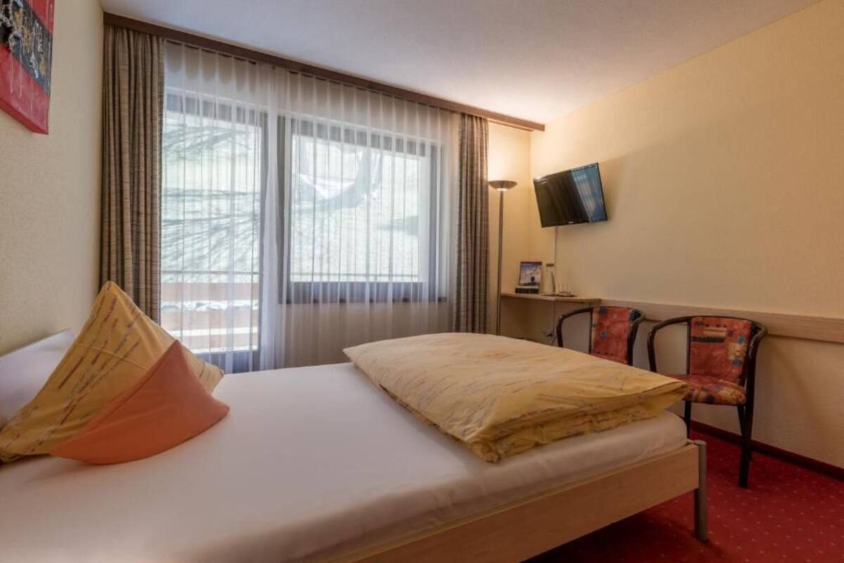 Habitación de hotel con cama, una silla, televisor en la pared y ventana con cortinas.