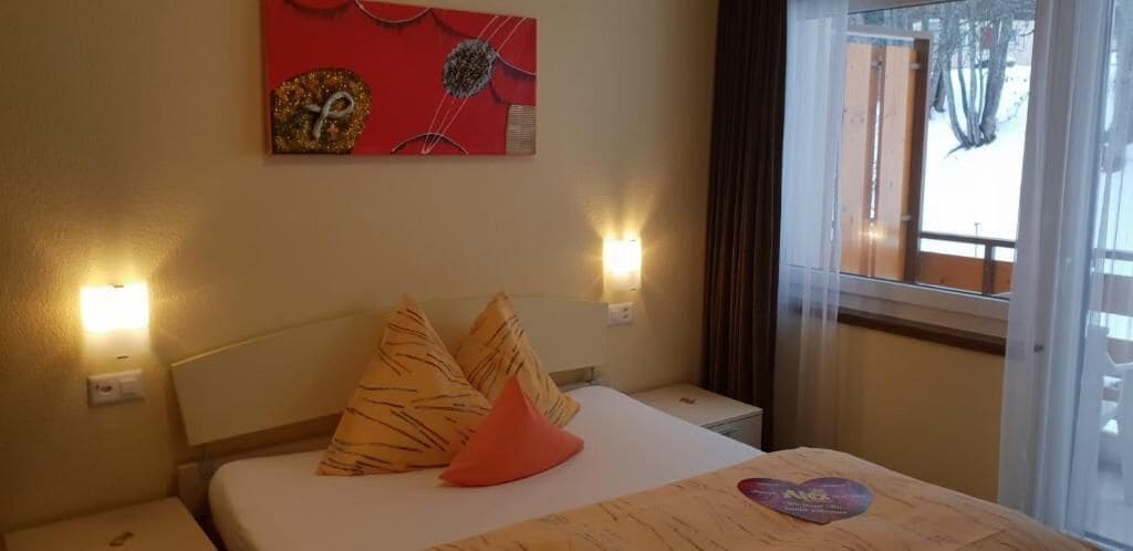 A hotel room with a bed, two pillows, a nightstand, a wall clock, and a window with curtains.