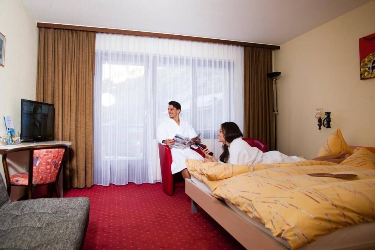 Habitación de hotel con cama, escritorio con silla, televisión y un gran ventanal con cortinas.