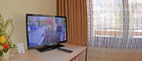 Una camera d'albergo con una televisione su una scrivania, un vaso con fiori e una finestra con tende.