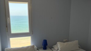 A hotel room with two beds, a window with a view of the sea, a nightstand with a lamp, and a teal bedspread.