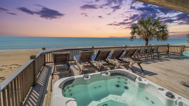 Een houten terras met ligstoelen en een jacuzzi met uitzicht op een strand bij zonsondergang.