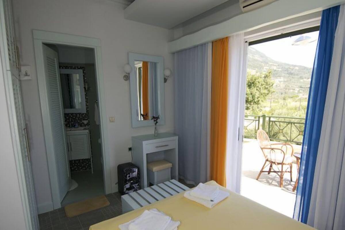 Habitación de hotel con cama, baño, lavamanos pequeño y balcón con una silla.