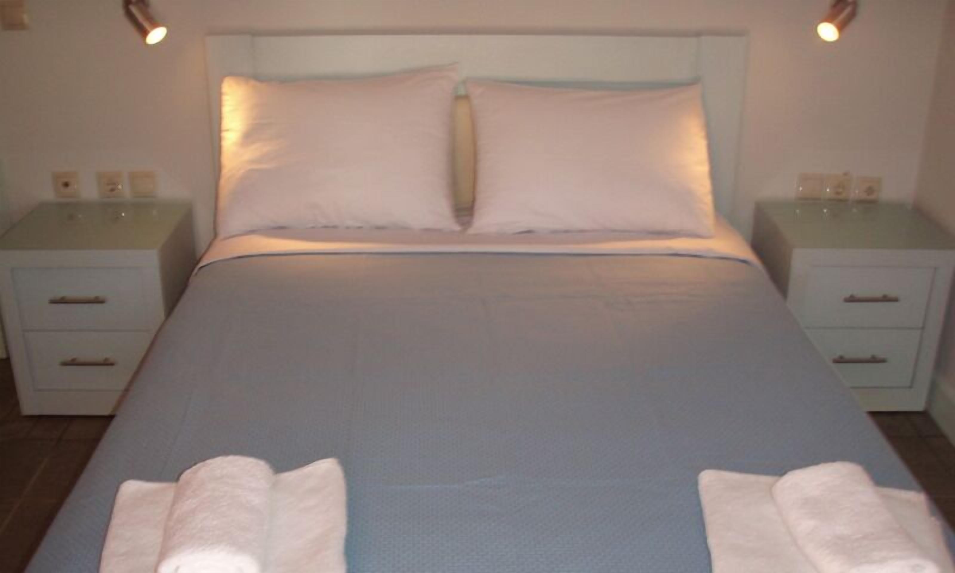 Un lit bien fait, avec du linge de lit blanc et deux oreillers, flanqué de deux tables de chevet sur lesquelles sont posées des lampes, dans une pièce simple et épurée.