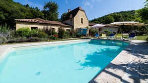 A swimming pool with lounge chairs and umbrellas, surrounded by a stone building and lush greenery. - Le Hibou Heureux (Castels et Bézenac)