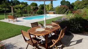 A wooden outdoor dining set with a large umbrella, a pool, and a garden in the background. - Le Hibou Heureux (Castels et Bézenac)