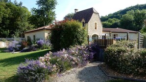 A stone house with a gravel pathway leading to a gated entrance, surrounded by lush greenery and flowering plants. - Le Hibou Heureux (Castels et Bézenac)
