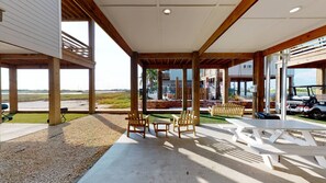 An open-air patio with wooden chairs and tables, a view of a golf course, and a modern building in the background. - PFC1006 Shared Pool, Ample Parking, Near Restaurants, Pet Friendly (Port Aransas)