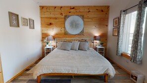 A bedroom with a wooden headboard, a large bed with striped bedding, two bedside tables with lamps, and a window with curtains. - PFC1006 Shared Pool, Ample Parking, Near Restaurants, Pet Friendly (Port Aransas)
