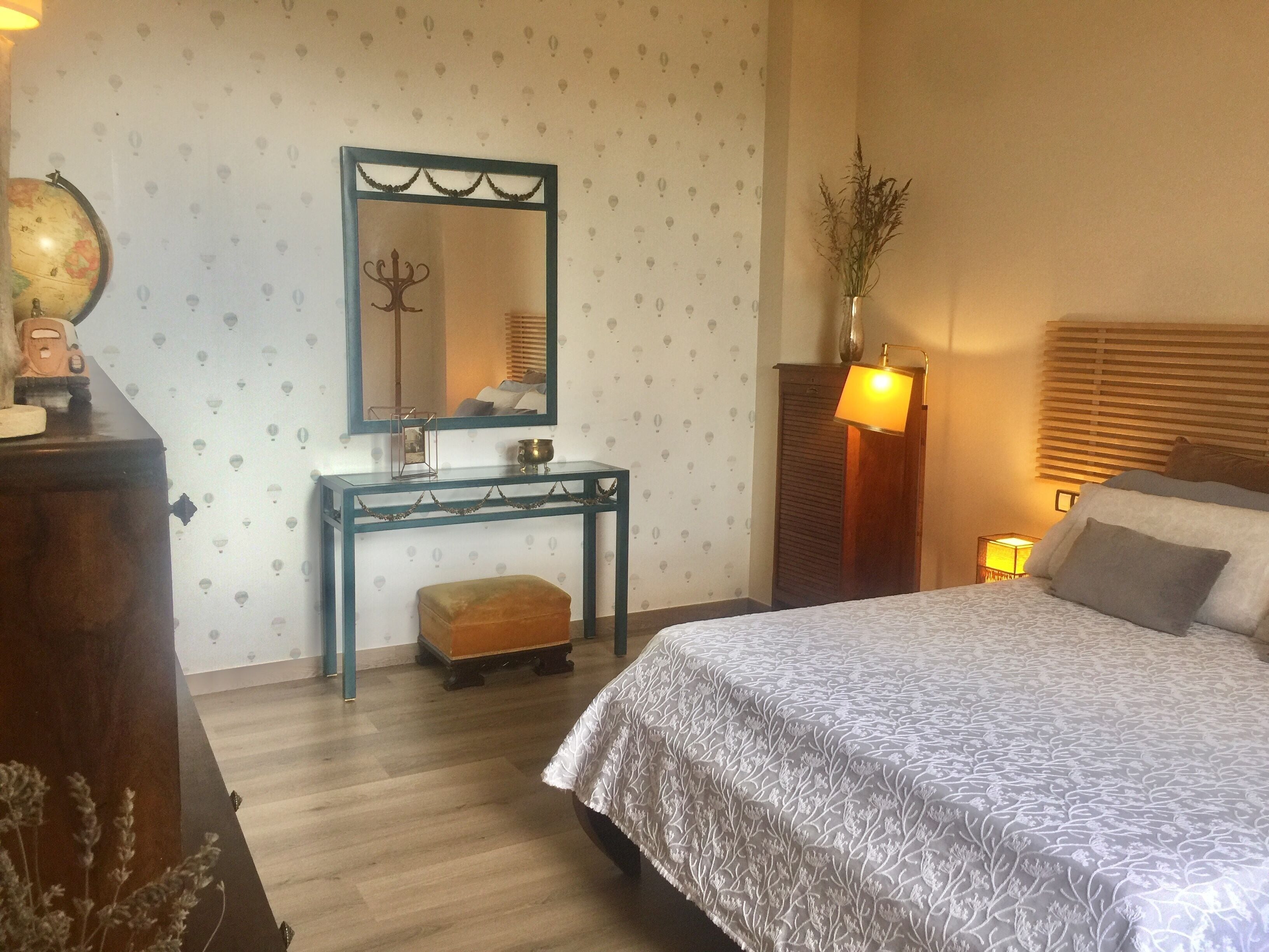A bedroom with a bed, a nightstand, a desk with a globe, a mirror, and a lamp.