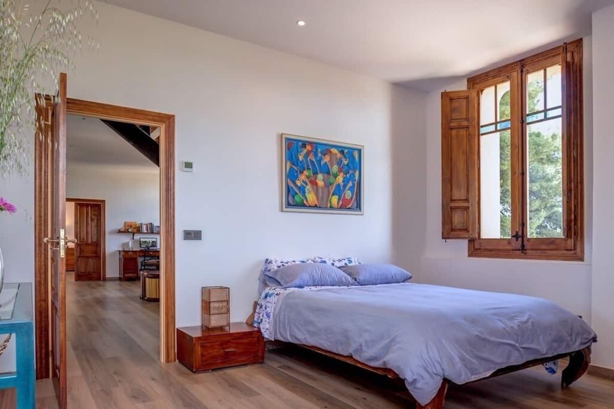 Une chambre avec un plancher en bois, un lit recouvert d’une courtepointe bleue, une fenêtre avec des volets en bois et un tableau accroché au mur.