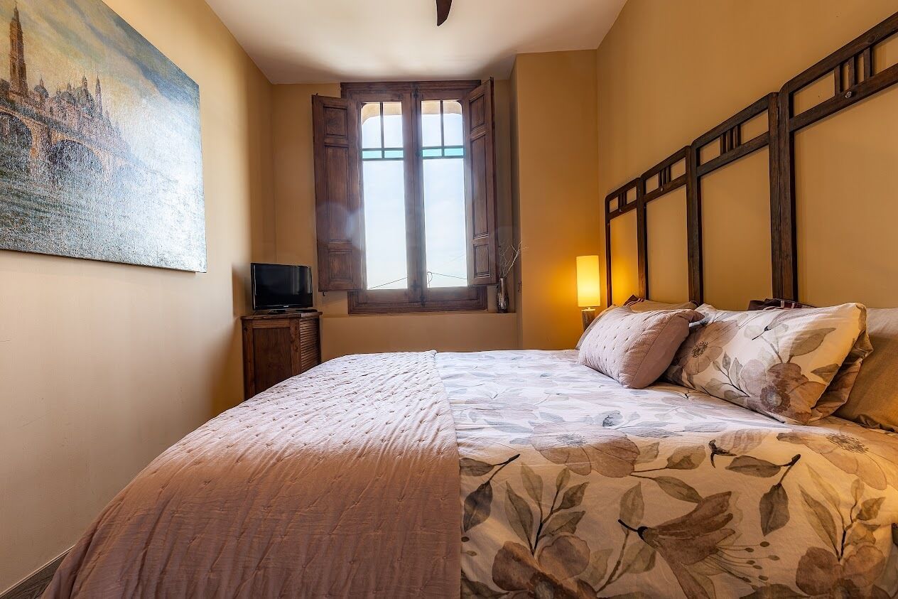 A bedroom with a bed, a painting, a window, and a nightstand.