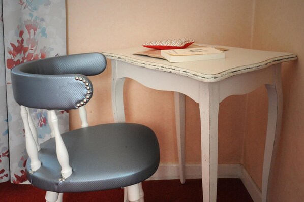 Una silla con asiento azul y patas blancas, una mesa blanca con un tazón rojo y papeles, y un papel tapiz floral de fondo.