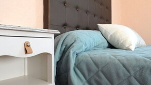 Una cama bien hecha con un cubrecolcha azul acolchado, una almohada blanca y una mesita de noche blanca con un cajón.