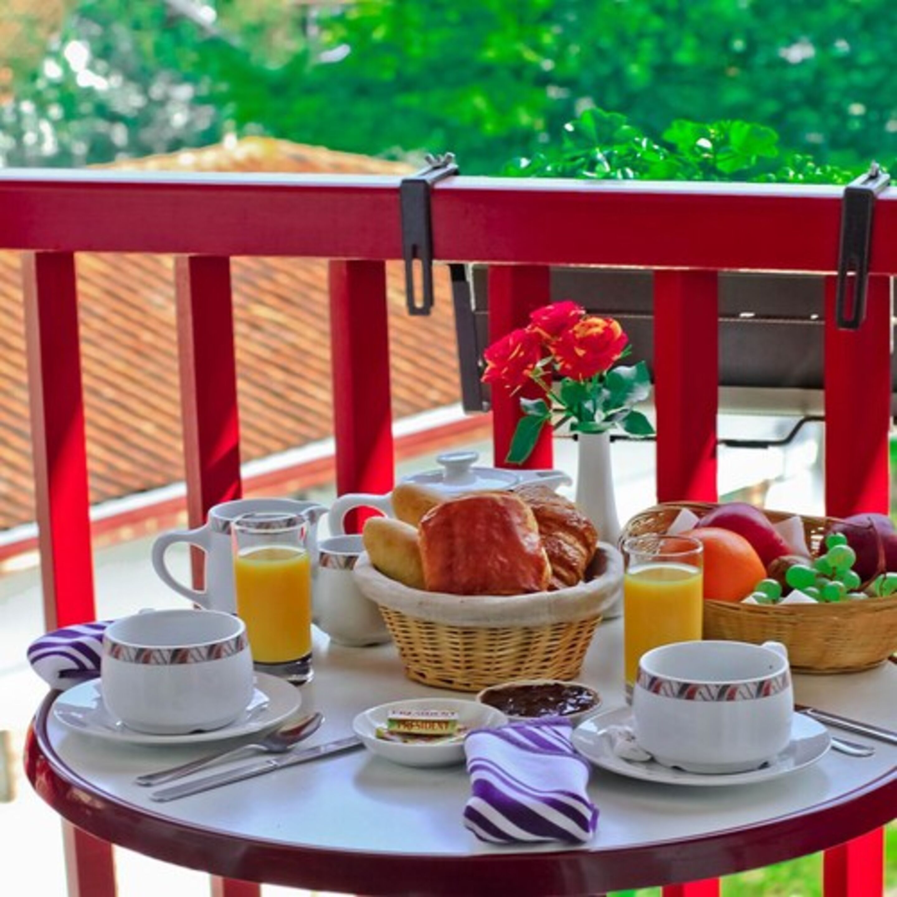 Desayuno en un balcón rojo con una cesta de mimbre, croissants, zumo de naranja y un jarrón con flores rojas.