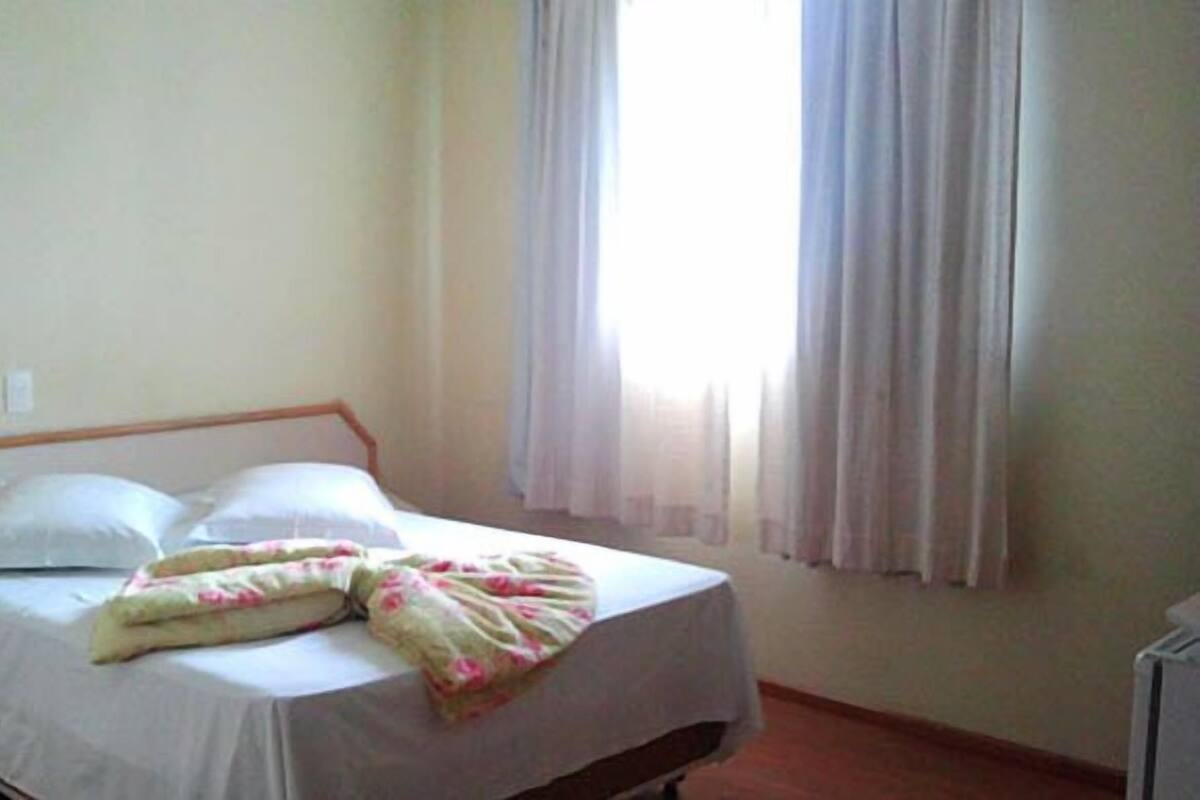 A single bed with white bedding and a floral-patterned blanket, a wooden headboard, and a window with sheer curtains.