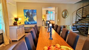 A dining area with a long wooden table, chairs, and a fruit basket. There is a painting on the wall, a staircase, and a mirror.