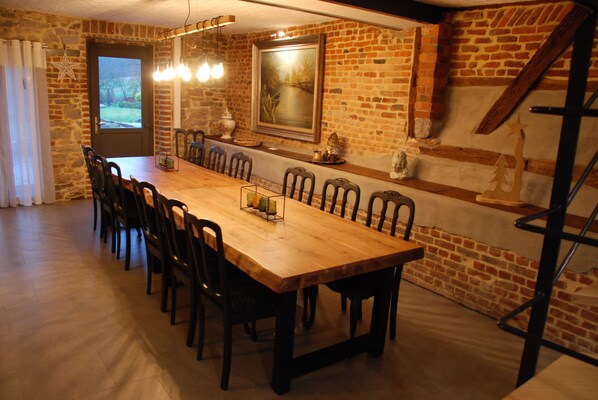 A long wooden dining table with chairs, a chandelier, and a painting on the brick wall.