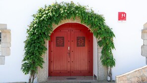 A red double door with decorative metalwork, surrounded by a stone wall and covered with green vines. - Relais Les Tilleuls (Hasparren)