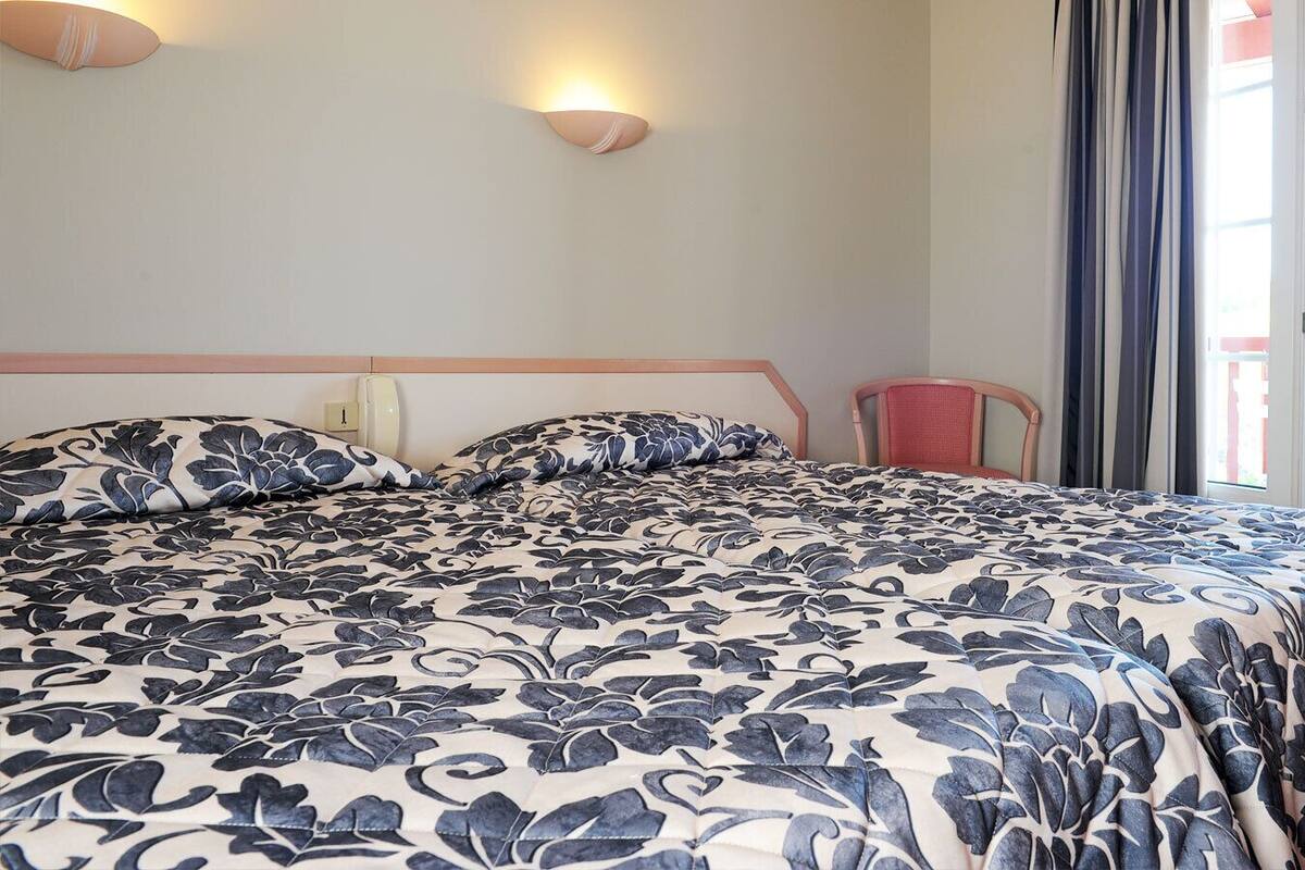 A bed with a floral bedspread, a chair, a wall-mounted light, and a window with curtains.
