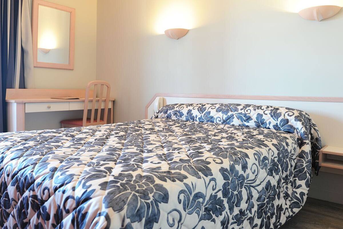 A hotel room with a bed, a desk, a chair, and a bedside table.