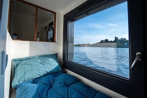 Una cabaña estrecha con una cama, una ventana que ofrece vistas a una masa de agua tranquila y una costa lejana, y un pequeño estante con objetos personales.