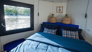 Quarto com uma janela grande, cama com roupa de cama azul, duas cestas trançadas na cabeceira e um quadro na parede.