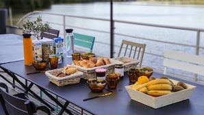 Mesa com itens de café da manhã, incluindo pão, frutas e bebidas, com vista para um lago.