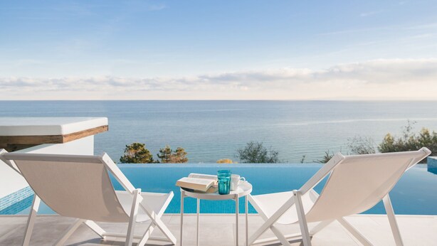 A poolside view with two white lounge chairs, a small table with a book and a glass, and a clear view of the sea. - Fiore Suites (Zakynthos)