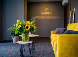 Quarto de hotel moderno com um sofá amarelo, uma mesinha redonda com um vaso de girassóis e uma parede com um desenho a tema Saturno.