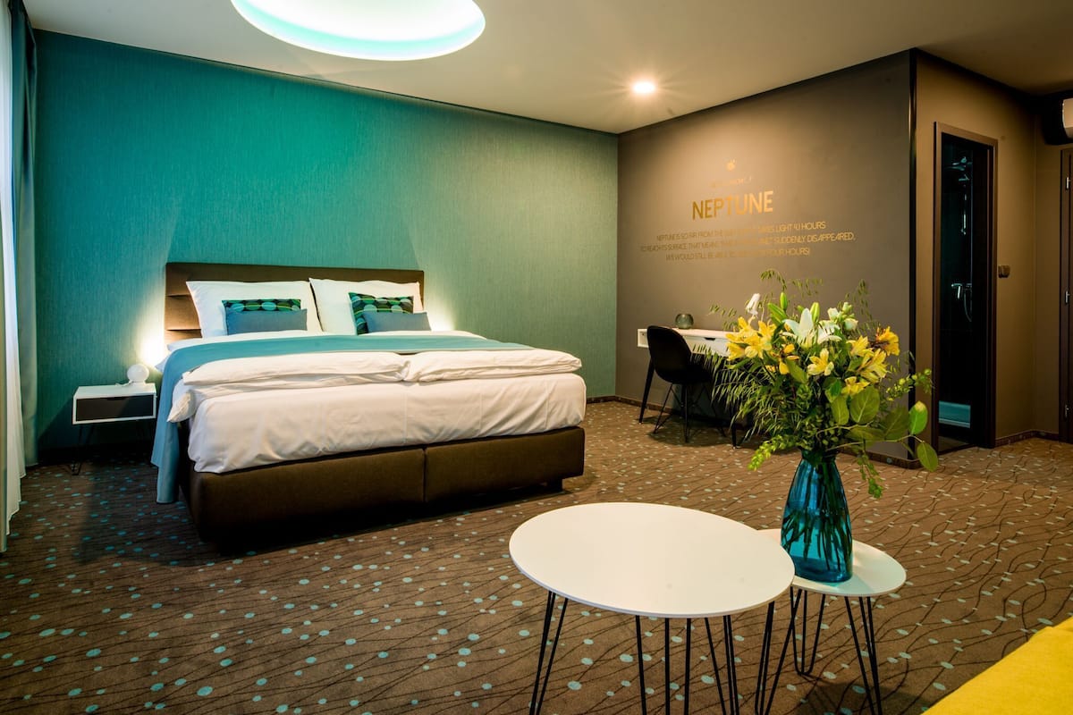 A hotel room with a bed, bedside tables, a desk, a chair, a small table, and a vase with flowers.
