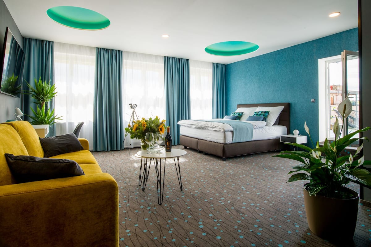 A hotel room with a bed, a yellow sofa, a coffee table with flowers, and a large window with curtains.