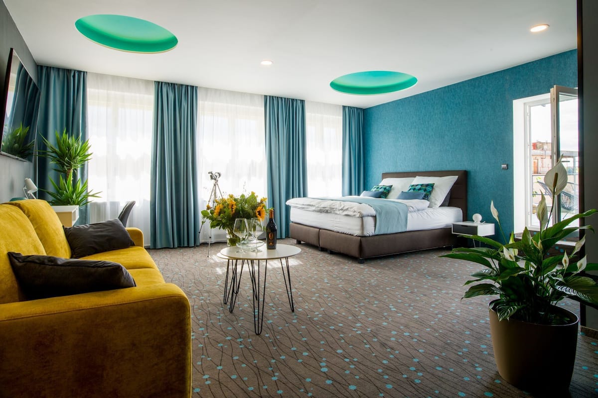 A hotel room with a bed, a yellow sofa, a round coffee table, and a large window with curtains.