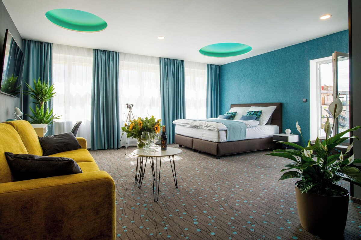 Una habitación de hotel con cama, sofá amarillo, mesa de centro con flores y un ventanal con cortinas.