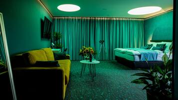 Ein Hotelzimmer mit einem gelben Sofa, einem Bett mit grüner Bettwäsche, einem kleinen runden Tisch mit Blumen und einem Stativ in der Ecke.