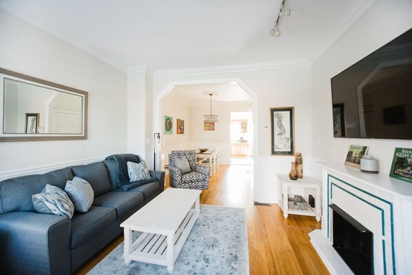 A living room with a grey sofa, a white coffee table, and a fireplace. There is a dining area in the background with patterned chairs and a chandelier.