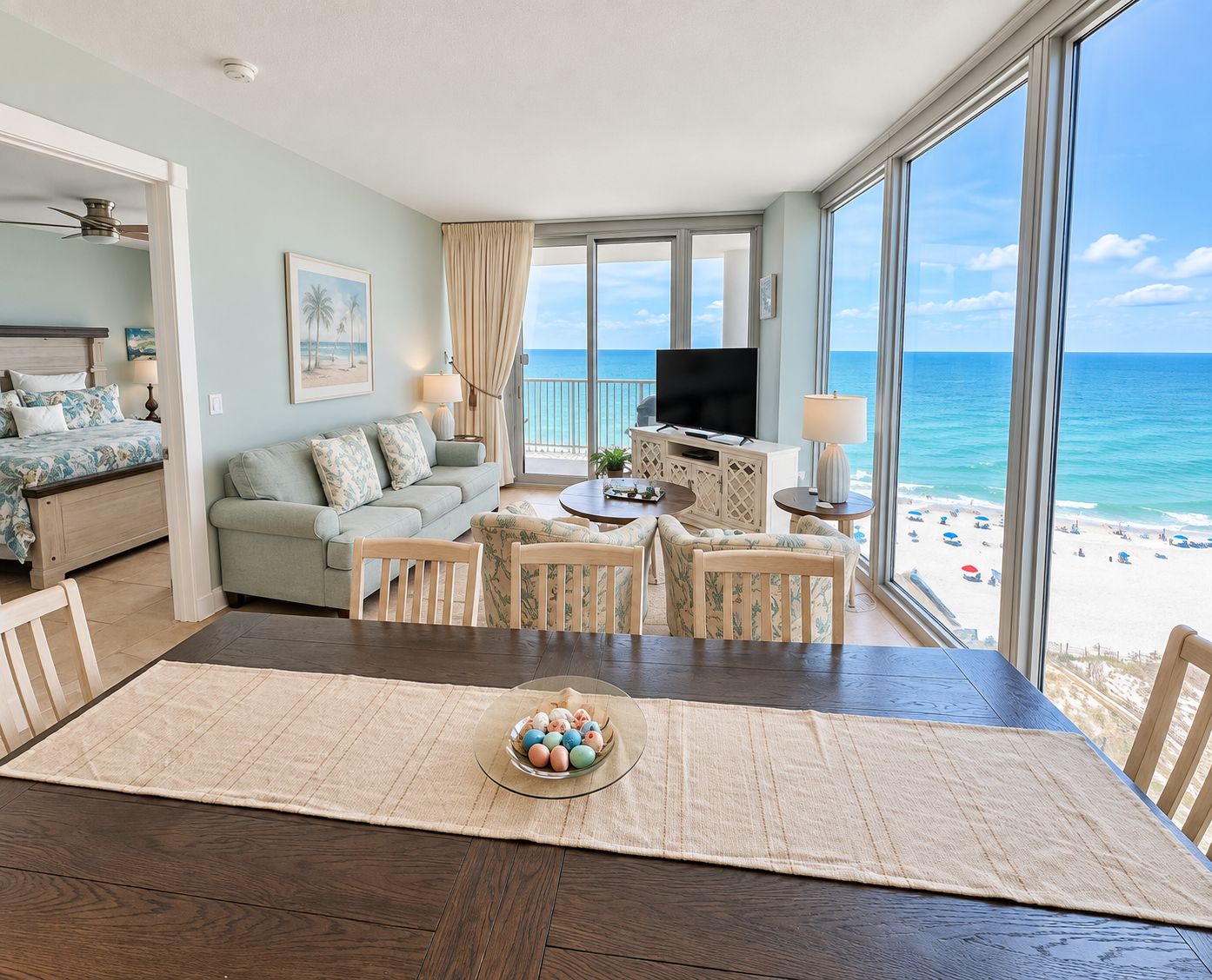 A beachfront hotel room with a balcony, a bed, a sofa, a TV, and a dining area with a table and chairs.