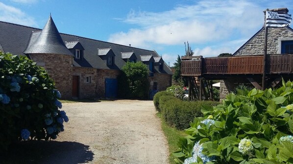 Un bâtiment en pierre surmonté d’un toit conique, doté d’une terrasse en bois et d’un chemin de gravier menant à une porte bleue.