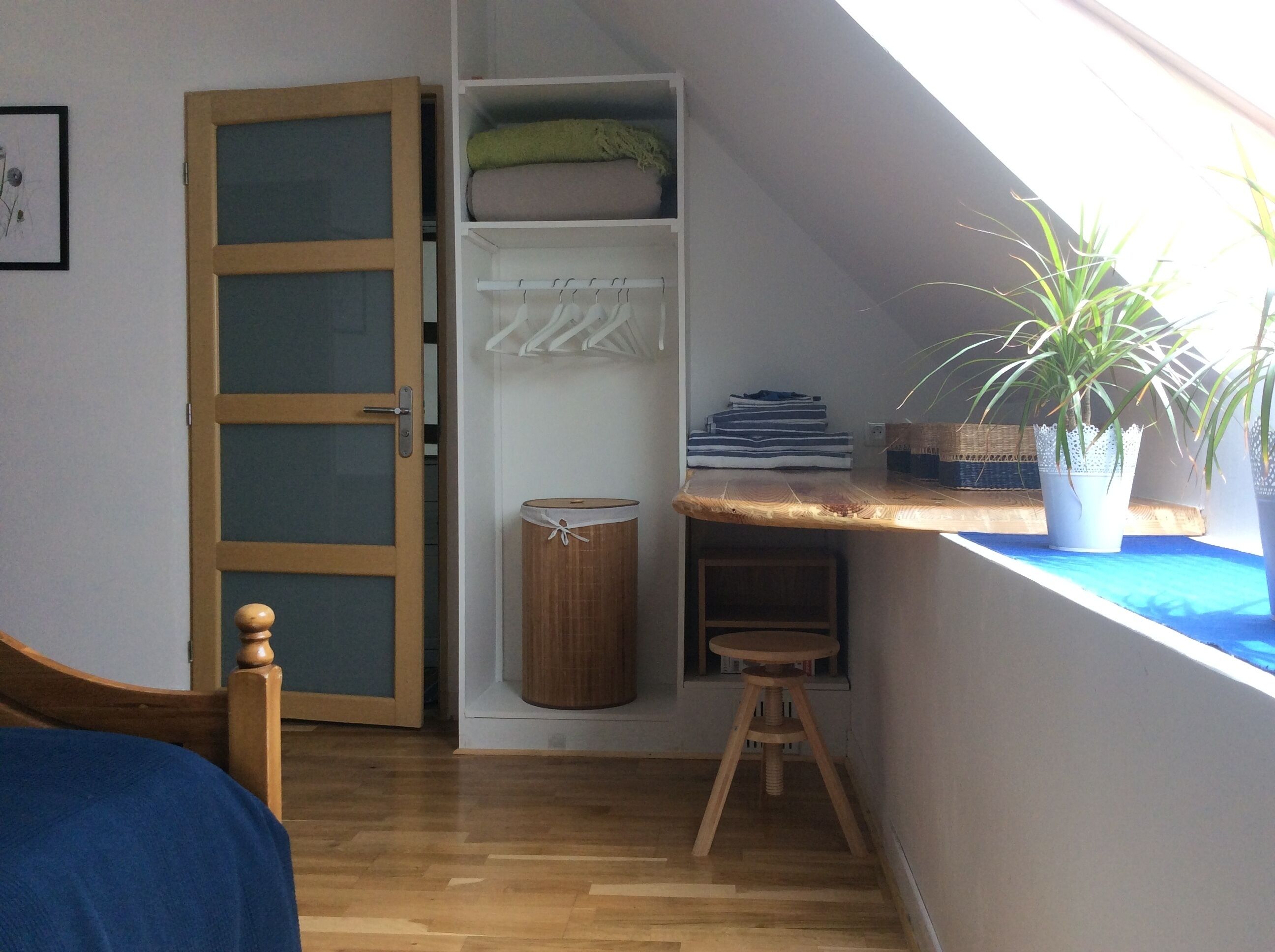A room with a sloped ceiling, a wooden bed, a wardrobe with a mirror, a small stool, and a window with a plant.