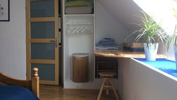 A room with a sloped ceiling, a wooden bed, a wardrobe with a mirror, a small stool, and a window with a plant.