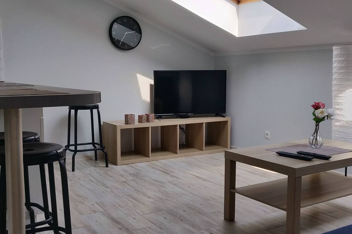 Een moderne keuken met een inbouwspoelbak, een eethoek met een tafel en krukken, een televisie op een houten standaard en een dakraam erboven.