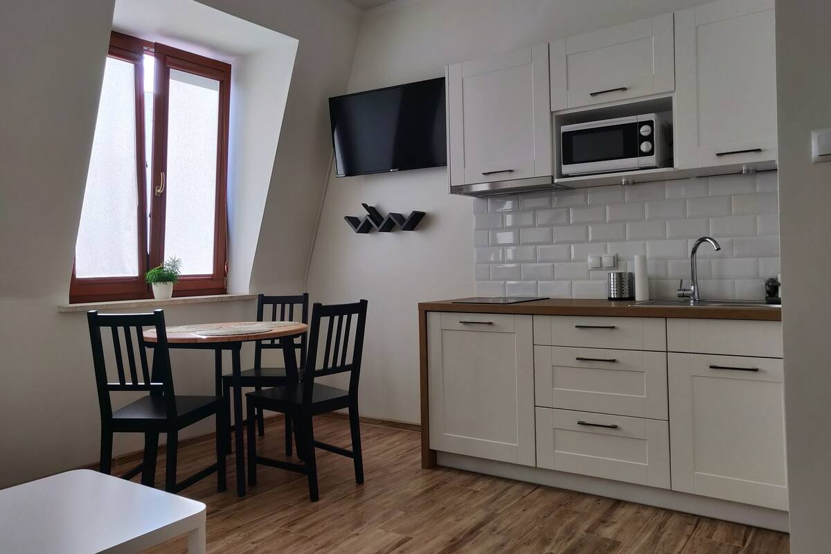 Een keuken met witte kastjes, een magnetron, een televisie en een raam met een plant op de vensterbank.