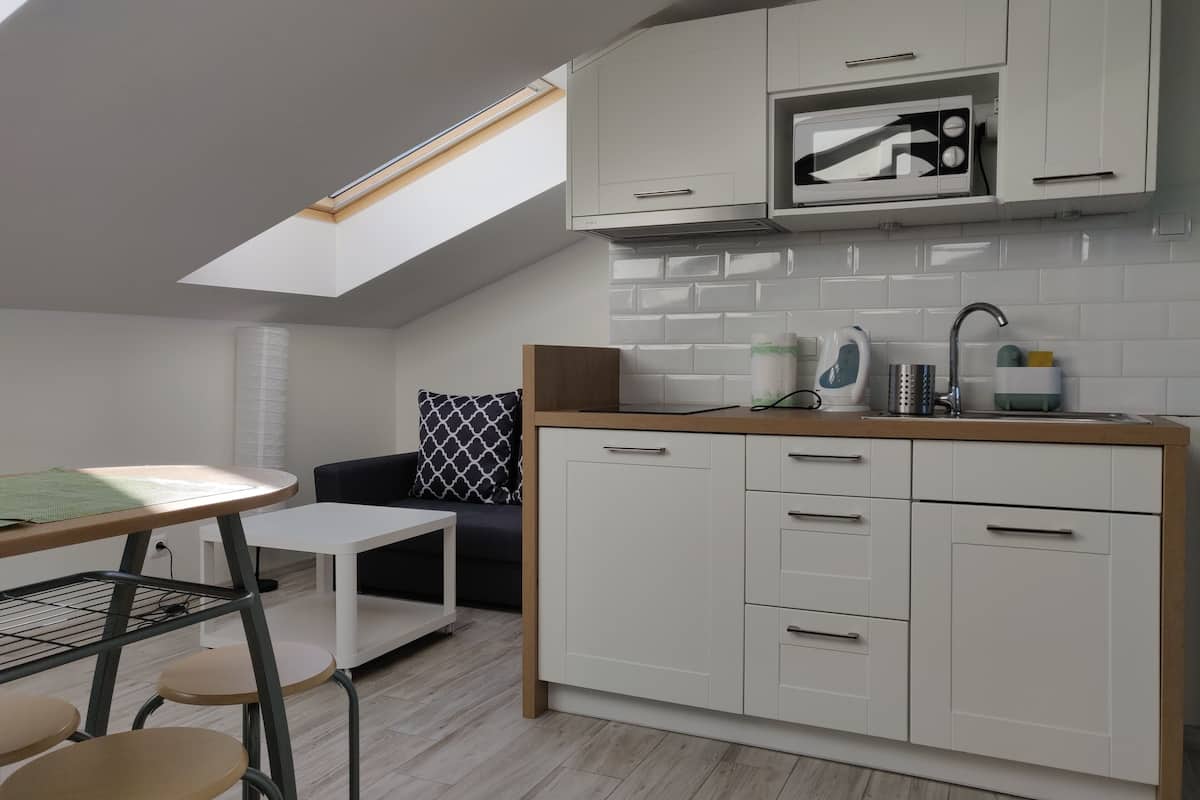 Een moderne keuken met witte kastjes, een magnetron en een spoelbak. Er staat een klein tafeltje met krukken en boven de keuken is een raam.