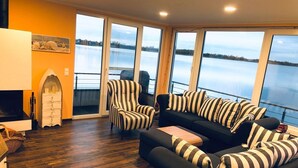 Un salón con un sofá negro y sillones a rayas, una mesa de centro de madera, y vistas a un lago a través de amplios ventanales.